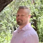 Christopher Landry, Owner and Clinician, wearing a light purple/pink long sleaves shirt with a canal featuring trees, grass an d green water , on the background